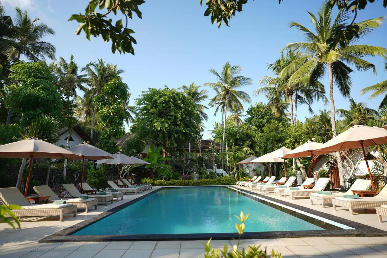 A long rectangular swimming pool is lined with white sun loungers and beige parasols, with palm trees and green foliage behind.