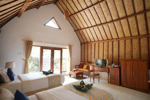 A bedroom with two beds under a thatched ceiling, a seating area, a desk with a television, and glass doors opening onto a garden with stone steps.