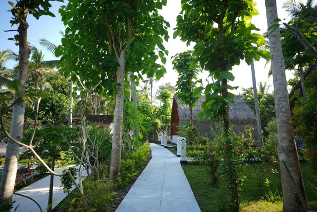 A pale concrete pathway runs between tall trees and shrubs toward a thatched-roof bungalow with white decorative posts.