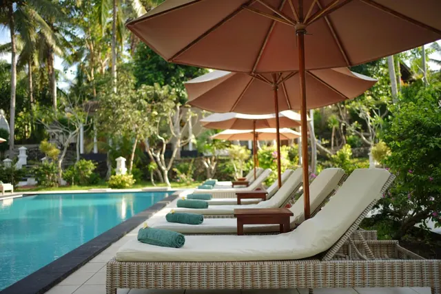 Sun loungers with cream cushions and rolled green towels sit beside a pool under tan parasols, with palm trees and greenery behind.