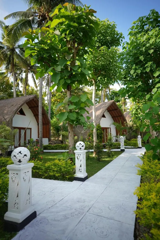 A paved pathway runs between white carved pillars and thatched-roof bungalows under tall palm and leafy trees.