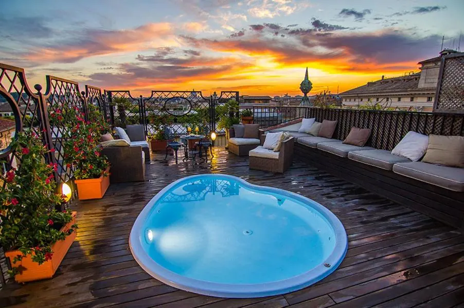 A rooftop terrace with a heart-shaped plunge pool, cushioned sofas and potted flowers, with rooftops visible under a sunset sky.