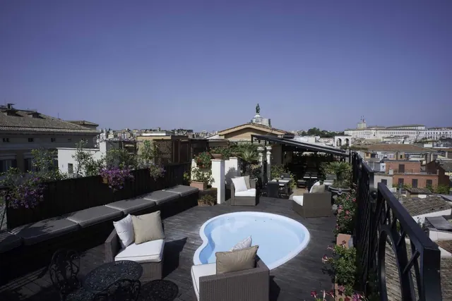 A rooftop terrace with a small kidney-shaped plunge pool, wicker seating and cushioned benches, overlooking the rooftops of the city under a clear sky.