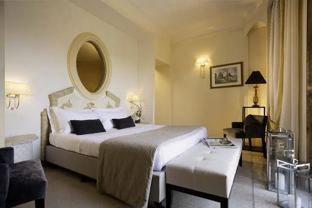 A bedroom with a made double bed under a round wall mirror, wall lamps on each side, a white bench at the foot of the bed, and dark armchairs in the corner.