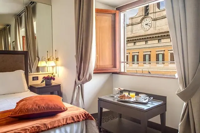 A bedroom with a made bed, a breakfast tray on a wooden table by the window, and a view of a building with a clock face.