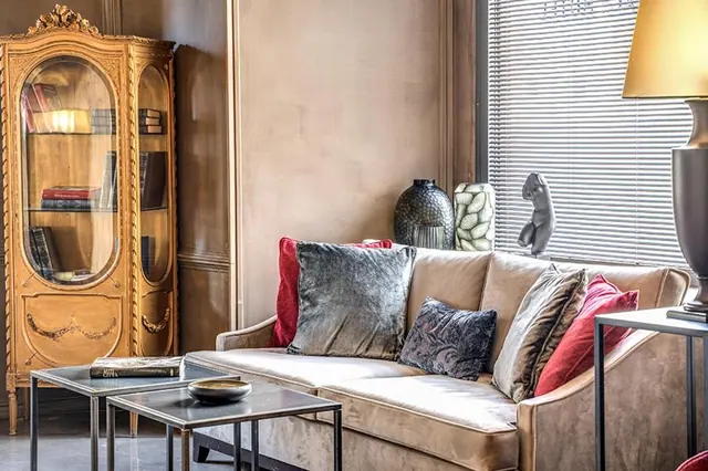 A living area with a beige velvet sofa and cushions, a glass-fronted display cabinet with books, metal side tables, and a window with blinds.