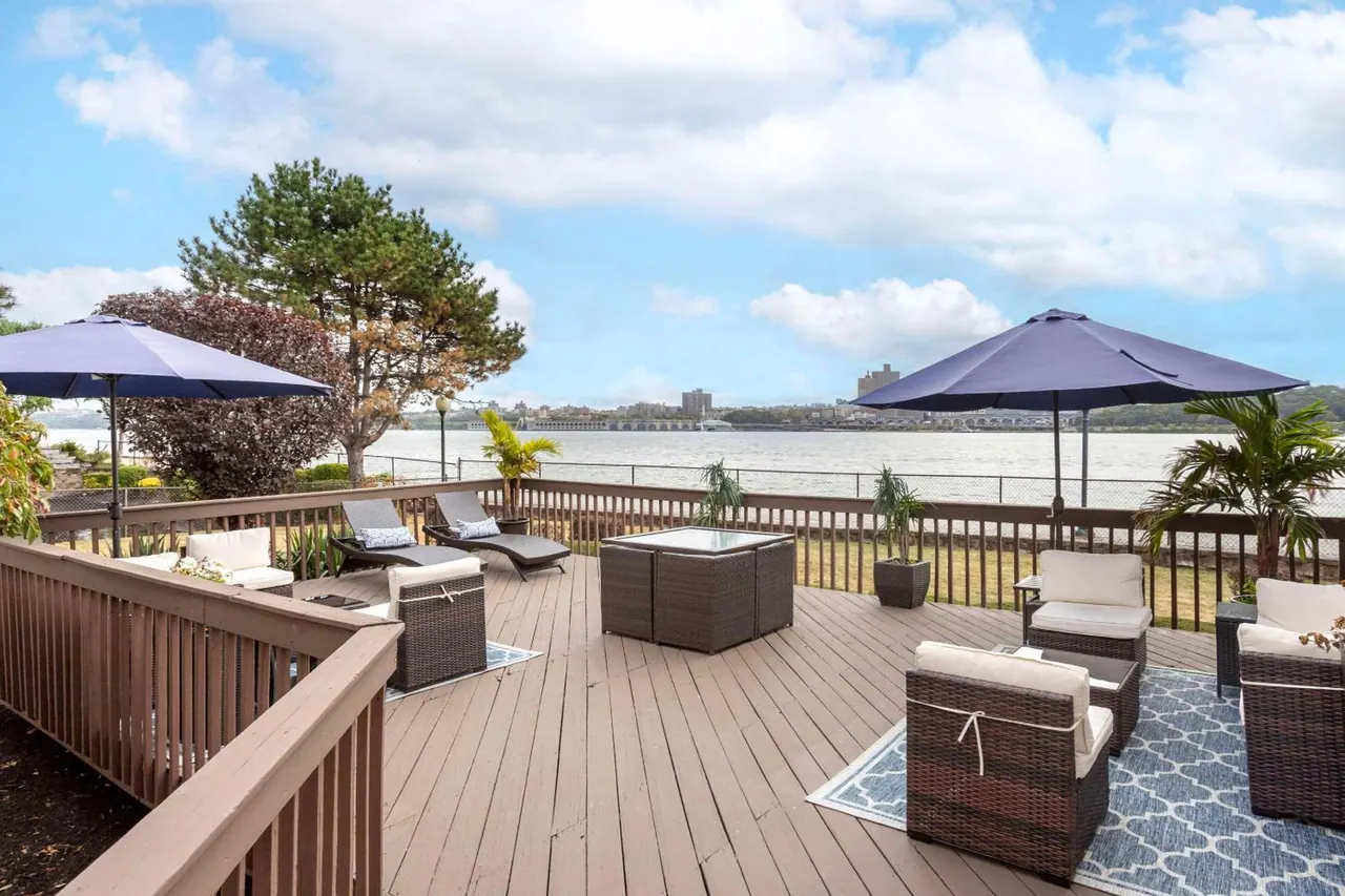 A wooden deck with wicker lounge furniture and blue umbrellas overlooks a wide river, with a city skyline on the far bank.