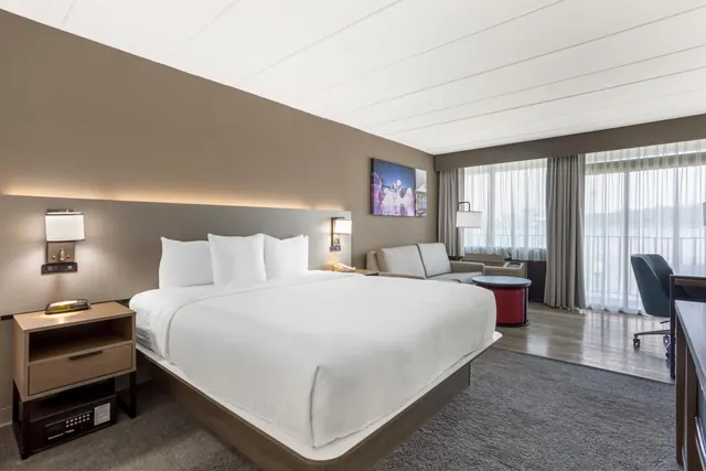 A king bed with white linens faces a seating area and floor-to-ceiling windows with sheer curtains that look out over water and trees.