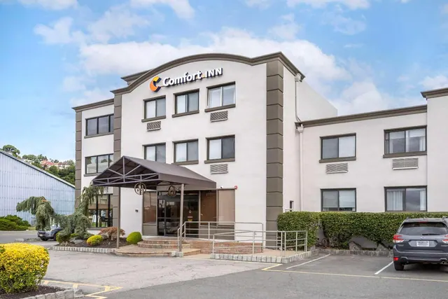 Exterior of a three-storey Comfort Inn with a covered entrance, metal railings at the front steps, hedges, and parked cars under a partly cloudy sky.
