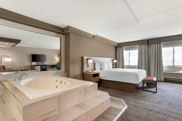 A bedroom with a king bed, a raised whirlpool bathtub in the foreground, and windows along the right wall.