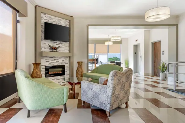 A lobby seating area with green sofas and patterned armchairs, a wall-mounted television above an electric fireplace, and lift doors on the right beside a checkered floor.