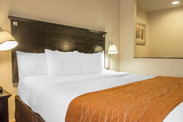 A king bed with white bedding, an orange throw and a dark wood headboard, with wall lamps on the wall beside it.
