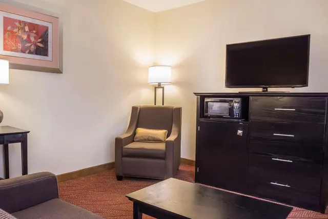 A living area with a upholstered armchair beside a lamp, a flat-screen television on a dark wooden dresser with a microwave, a coffee table, and a framed abstract floral print on the wall.