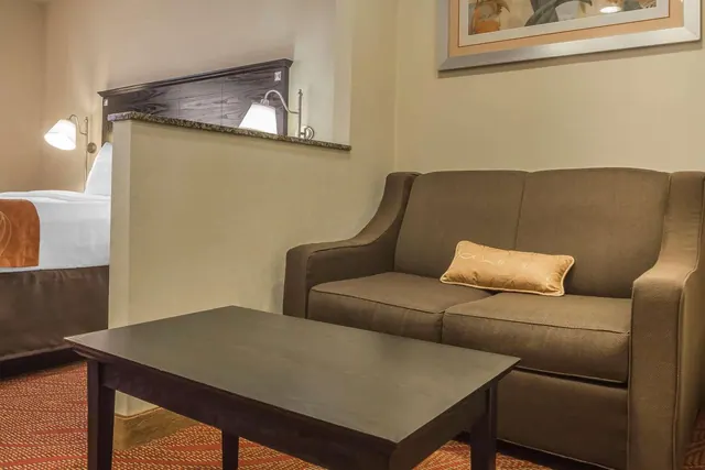 A brown loveseat with a gold cushion and a dark wooden coffee table sit beside a partially visible bed with wall lamps in a hotel room.