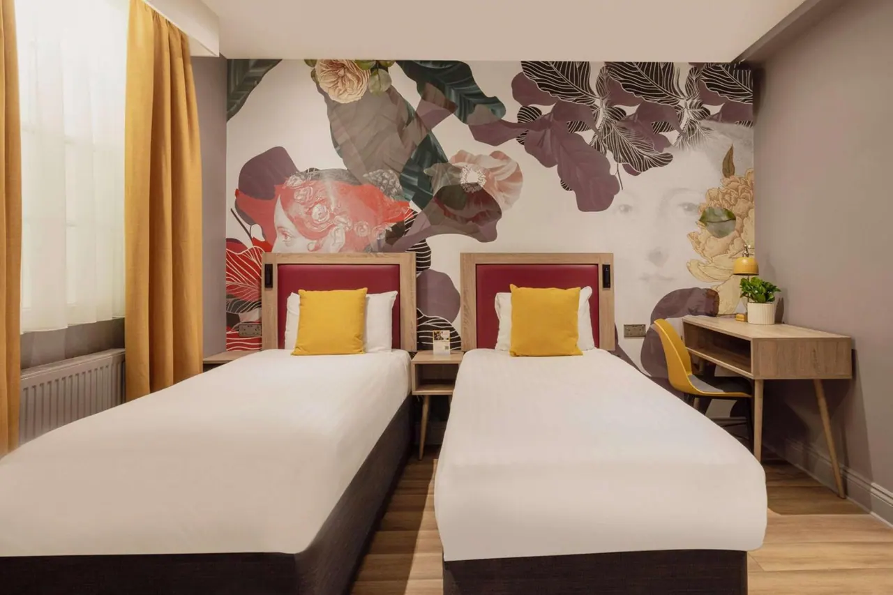 A bedroom with two single beds with red headboards and yellow cushions, a floral wall mural, and a wooden desk with a yellow chair.