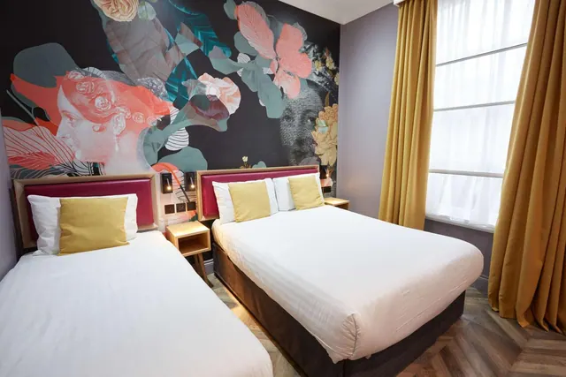A hotel bedroom with a single bed and a double bed in front of a dark floral wall mural, with yellow curtains at the window.