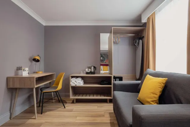 A hotel room with a wooden desk and yellow chair, a grey sofa with a yellow cushion, a shelf holding rolled towels and a kettle, and a wardrobe with an ironing board.