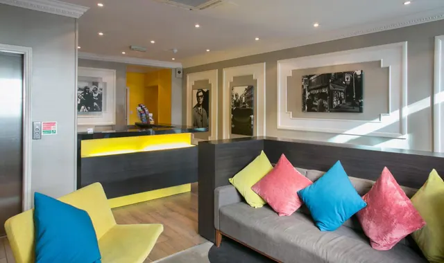 A lobby with a yellow-lit reception desk, a grey sofa with coloured cushions, a yellow armchair, black and white framed photographs on the wall and a lift on the left.