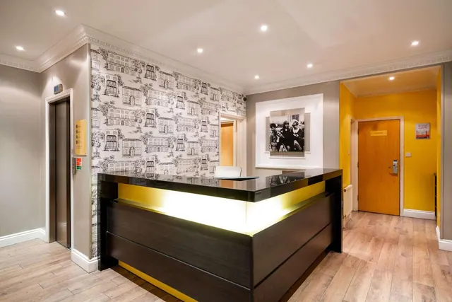 A dark reception desk with a backlit yellow panel stands in a lobby with patterned black-and-white wallpaper, a lift on the left and a corridor with a wooden door on the right.