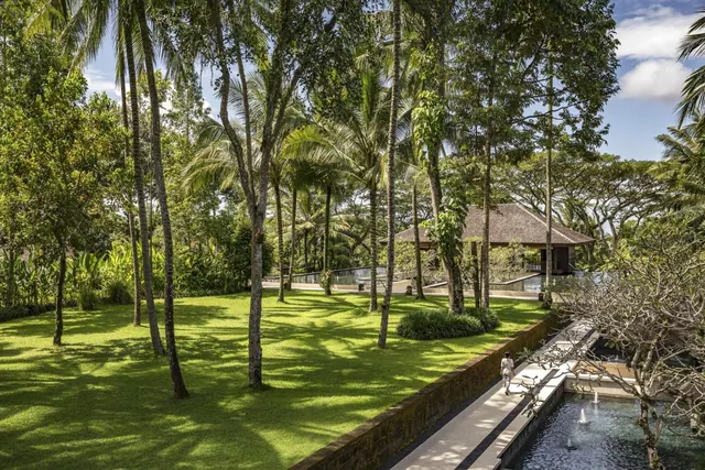 A manicured lawn with palm trees, a thatched pavilion and a narrow fountain pool, with one person walking along a stone path.
