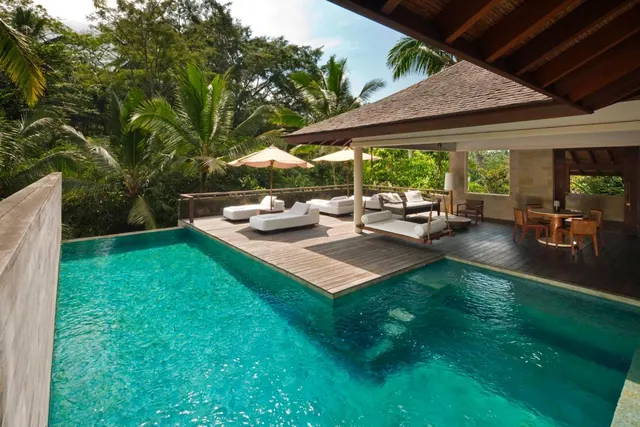 A private swimming pool sits beside a timber deck with sun loungers, parasol umbrellas, a swing bed and a covered dining area, with jungle trees behind.