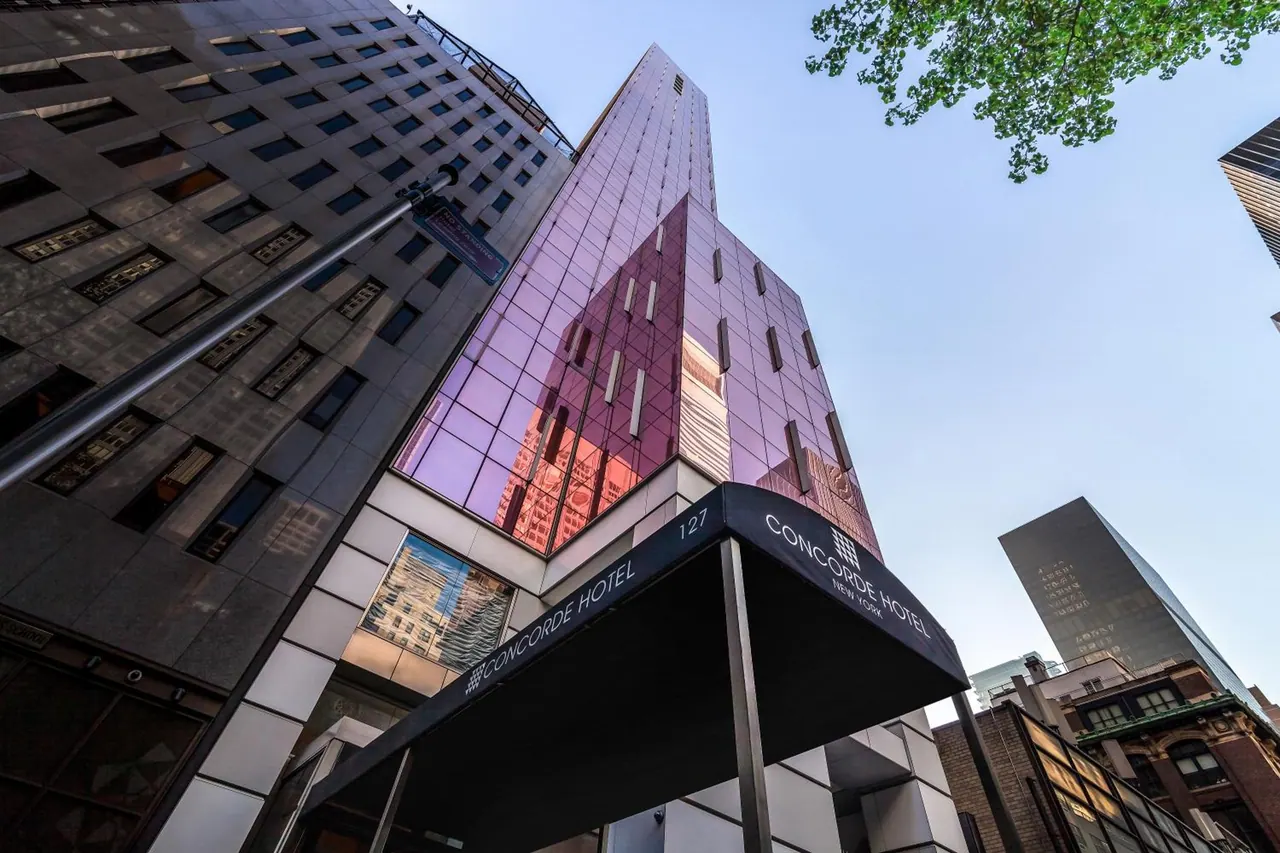 Low-angle view up the Concorde Hotel New York building, showing its black entrance awning and tall tower with pinkish reflective glass.