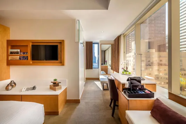 A hotel room with a bed in the foreground, a wall-mounted television, a desk with a coffee machine, a seating bench, and floor-to-ceiling windows looking onto city buildings.