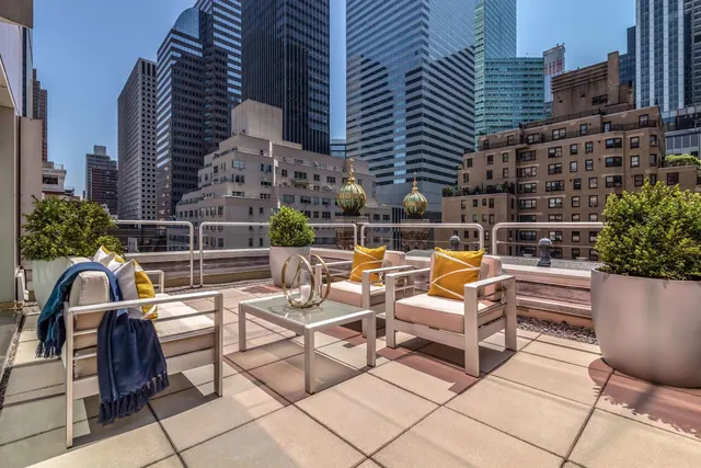 A rooftop terrace with white lounge chairs, yellow cushions, a glass coffee table and potted shrubs, overlooking a dense cluster of skyscrapers and midrise buildings under a clear sky.