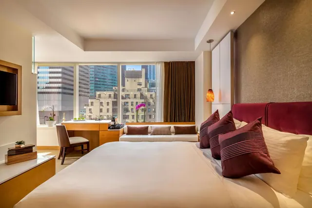 A bedroom with a large bed with red and cream pillows, a wooden desk and chair, a window bench, and a view of city buildings through floor-to-ceiling windows.