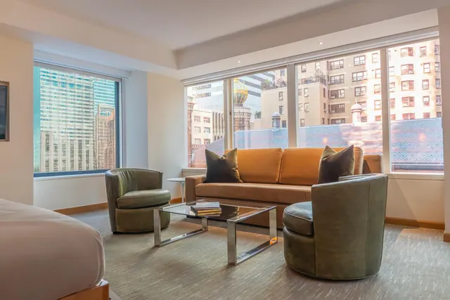 A hotel room seating area with an orange sofa, two green armchairs, a glass coffee table and large windows looking onto city buildings.