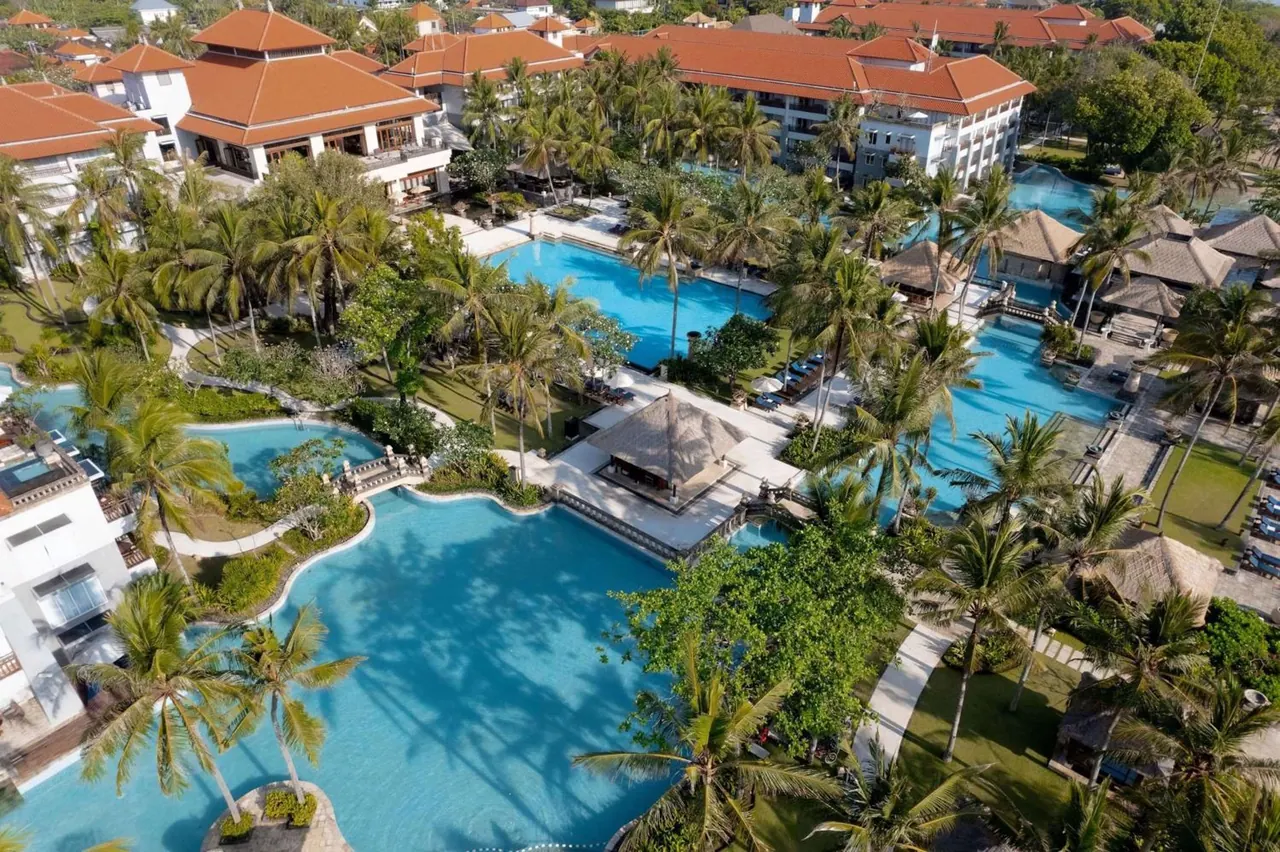 Aerial view of a resort with several swimming pools, palm trees, thatched pavilions and orange-roofed buildings.