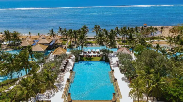 Aerial view of a resort with a large rectangular pool, rows of sun loungers and white umbrellas, palm trees, thatched-roof pavilions and the ocean beyond.