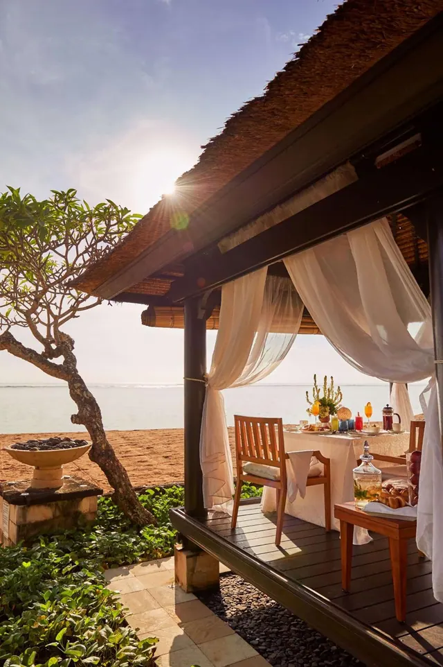 A thatched beachside pavilion with a set breakfast table and white curtains overlooks the sea and sandy shore.