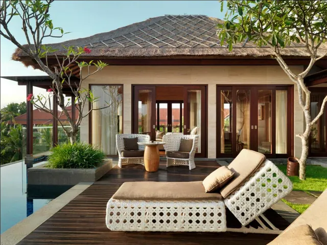 A timber deck terrace with white wicker loungers and armchairs sits in front of glass doors of a thatched-roof villa, with an infinity-edge pool at the left edge of the frame.