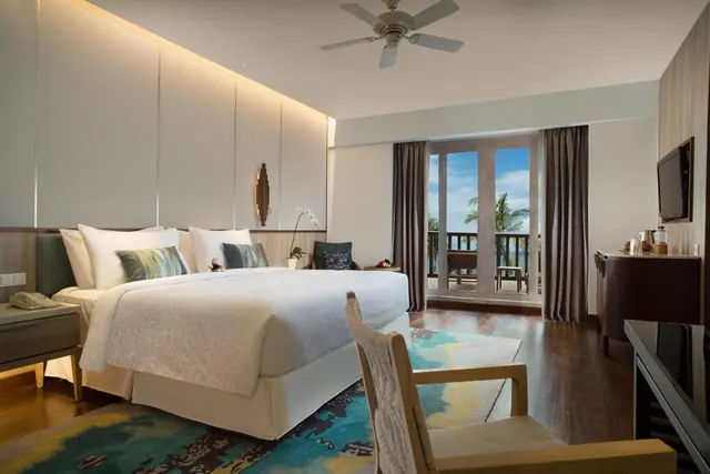 A bedroom with a king bed, ceiling fan, wall-mounted television and glass doors opening onto a balcony with a view of the sea and palm trees.