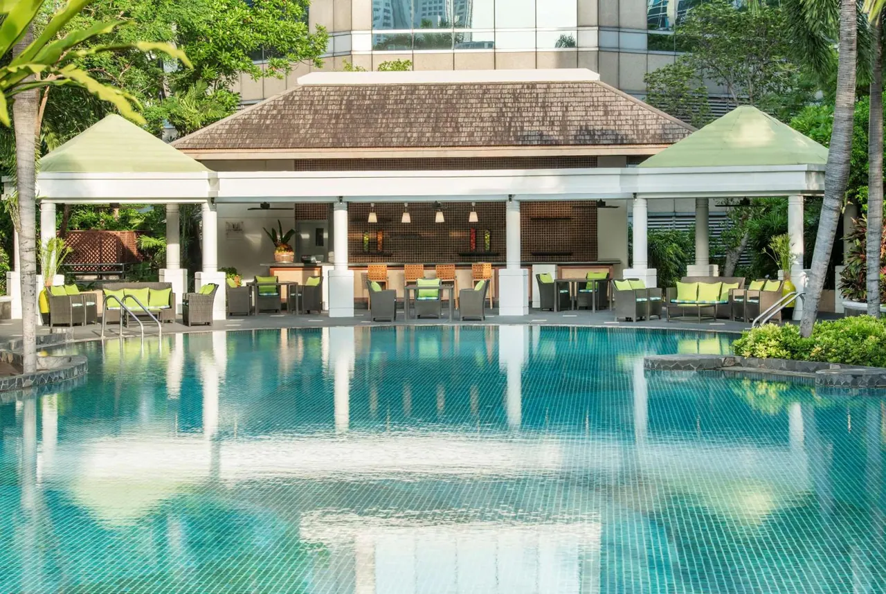 A large tiled swimming pool sits in front of an open-sided poolside bar pavilion with green cushioned seating, flanked by palm trees.