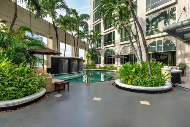 A paved poolside terrace with a small pool, a wall-mounted water feature, palm trees, brown shade umbrellas and a rattan seating area beside a tiled hotel building.