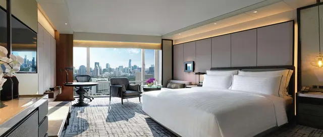 A bedroom with a large king bed, a round work desk and armchair, and floor-to-ceiling windows overlooking a city skyline.