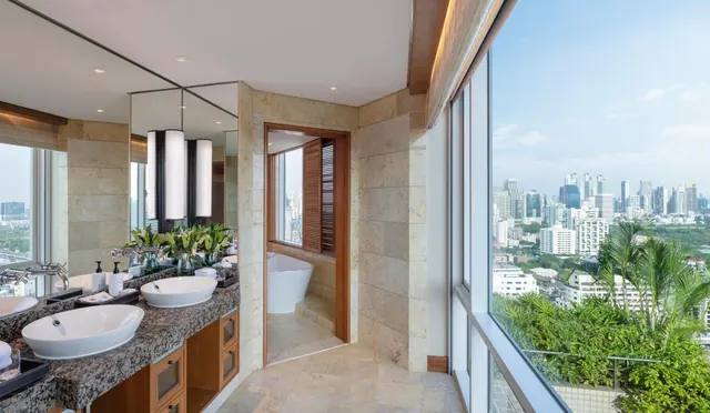 A bathroom with a double vanity and mirrors, a freestanding bathtub visible through the doorway, and floor-to-ceiling windows looking out over a city skyline.