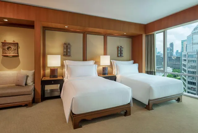 A hotel bedroom with two made-up twin beds, a sofa on the left, and floor-to-ceiling windows overlooking a city skyline.