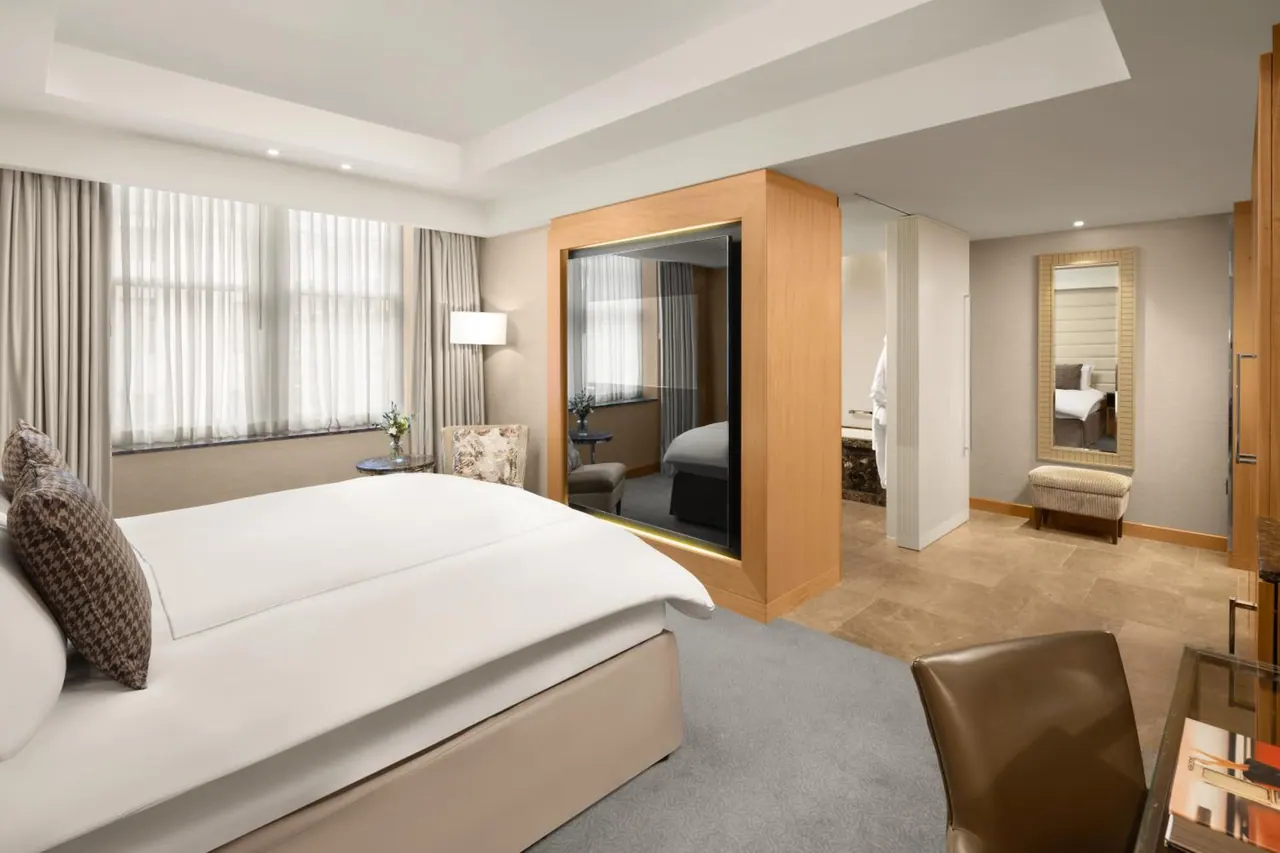 A bedroom with a made king bed, an armchair by the window, a wooden wardrobe, and a framed mirror opening into an adjoining room.
