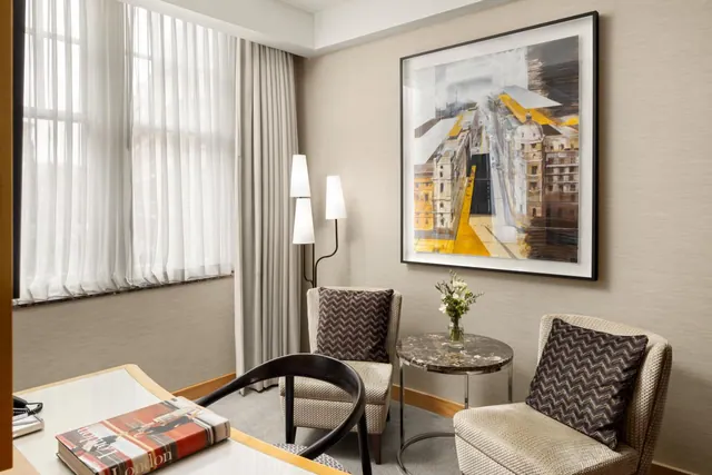 A hotel room seating area with two upholstered armchairs, a round marble side table with flowers, a floor lamp, a large framed city artwork, and a desk with a London book on it.