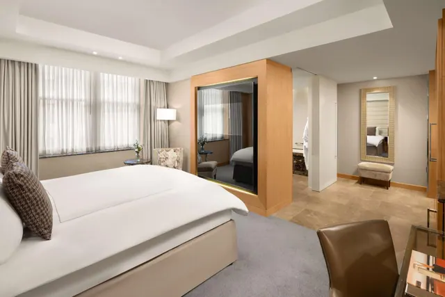A king bed with white linen and patterned cushions sits beside a sheer-curtained window, with an armchair, a mirrored wooden partition, and a bathroom visible in the background.