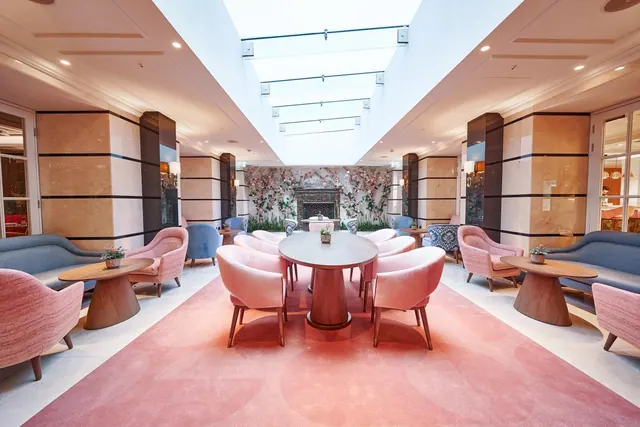 A lobby seating area with pink armchairs and round tables on a pink rug beneath a glass skylight, with a floral wall and fireplace in the background.