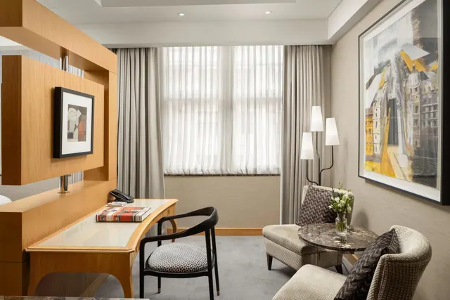 A hotel room with a wooden desk and black chair, a marble side table with two armchairs, floor lamps, and a large painting beside a sheer-curtained window.
