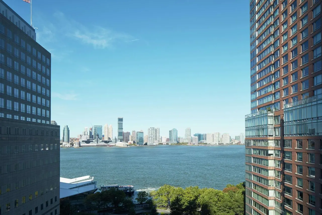 View past two high-rise buildings over a river to a distant city skyline, with trees and a moored boat in the foreground.