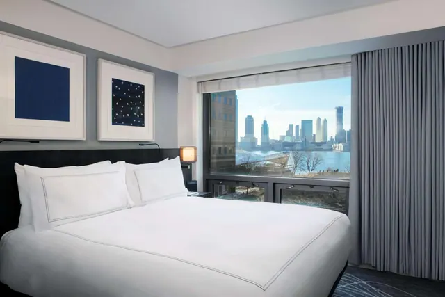 A bedroom with a made king bed, framed artwork, a bedside lamp, and a large window looking over water toward a city skyline.