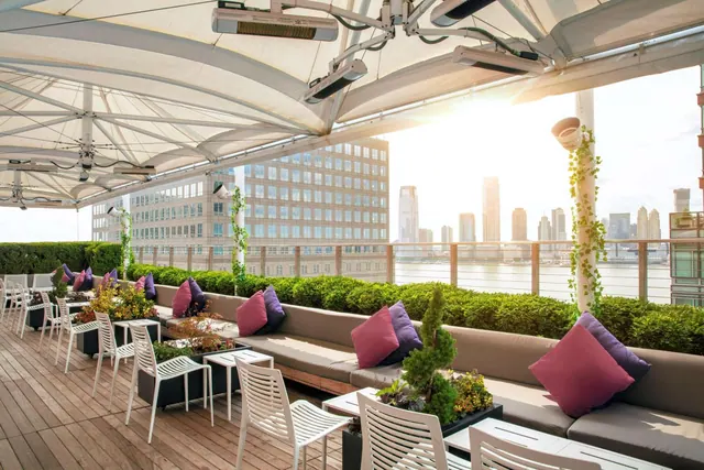 A rooftop terrace with white chairs, planters and cushioned banquette seating overlooks a river and a city skyline under a canopy with overhead heaters.