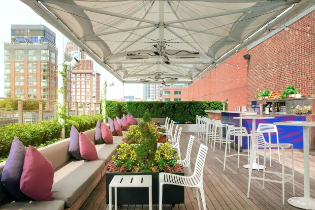 A rooftop terrace under a large canopy, with built-in bench seating with purple and pink cushions, white high chairs and tables, a bar counter, and city buildings visible beyond the edge.