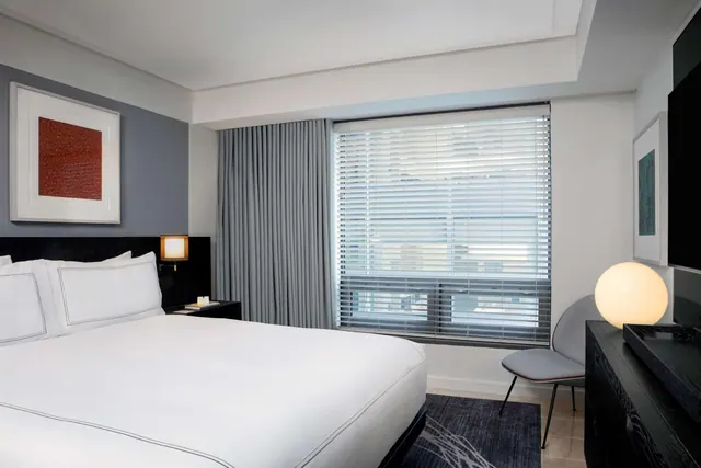 A bedroom with a made king bed, closed window blinds and grey curtains, a round table lamp, an armchair and a wall-mounted television.